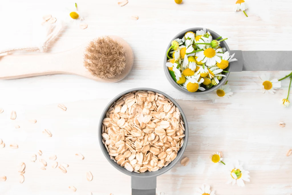A brush, a measuring cup with oats, and chamomile flowers.