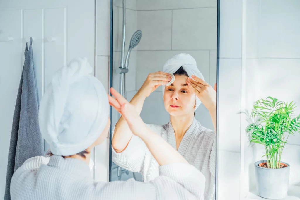 A female cleaning her forehead with a cotton pad in front of the bathroom mirrior. 